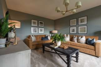 A plush living room features a dark wood coffee table with a fern centerpiece in the foreground, while the background showcases two tan sofas against a muted green wall adorned with framed artwork.
