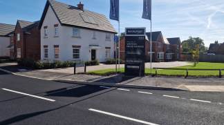A paved road with white lines in the foreground leads to a development of new houses with a sales sign in the background.