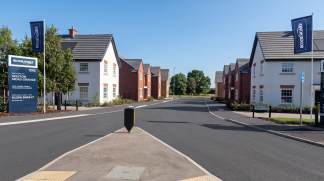 A paved road leads through a new housing development with modern houses on either side, framed by trees and a clear blue sky in the background.