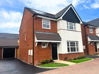 A modern red brick house with white trim and a black front door sits in the foreground, while a row of similar houses and a cloudy blue sky form the background.