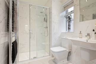 A modern bathroom features a glass-enclosed shower in the foreground and a toilet and sink with a window in the background, all adorned with white marble-effect tiles.