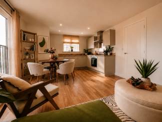 A modern living and dining area with a kitchen in the background features a plush green ottoman and armchair in the foreground, a round dining table with chairs, and a sleek kitchen with light cabinetry.