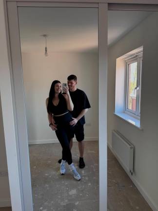 A couple in athletic wear poses for a mirror selfie in a room with bare walls and a window, reflecting the unfinished state of the space.