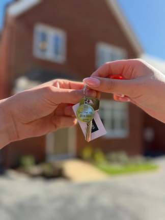 Hands exchange a set of keys with a decorative fob in front of a blurred brick house.