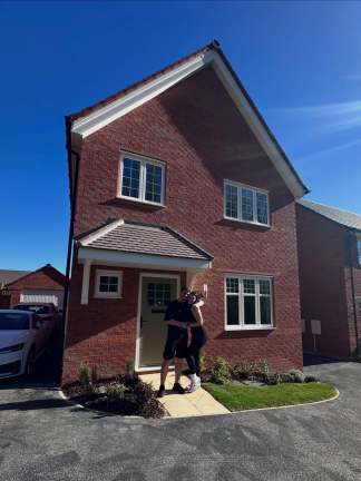 A happy couple embraces on the paved walkway in front of their new red brick house, with a clear blue sky overhead and other houses visible in the background.