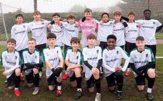 A youth soccer team poses for a photo in their white and green uniforms on a muddy field with a soccer net in the background.
