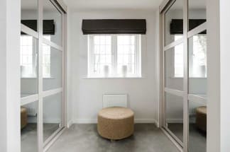 A spacious, well-lit room with large windows, a plush ottoman in the foreground, and mirrored closet doors reflecting the natural light.