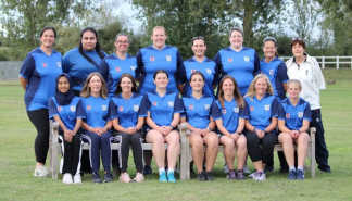 A group of women in blue cricket uniforms pose for a photo on a grassy field with a white fence and trees in the background.