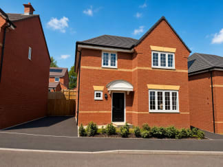 A modern red brick house with a black door and white-framed windows sits on a paved driveway, with other houses and a blue sky in the background.