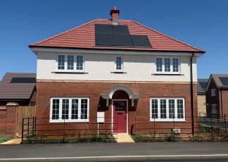 A modern two-story house with a red-tiled roof and solar panels sits behind a black metal fence, with other similar houses visible in the background under a clear blue sky.
