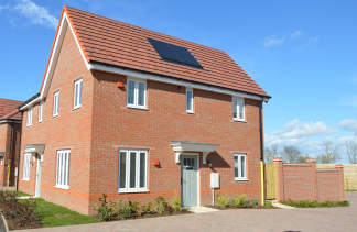 A modern brick house with white-framed windows and solar panels on its red-tiled roof stands on a paved driveway, with a wooden fence and distant trees under a clear blue sky.