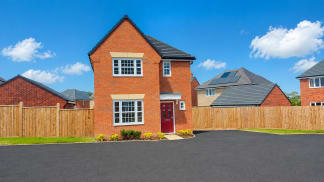 A new brick house with a red door stands in the foreground, with a wooden fence and a paved driveway, against a bright blue sky with scattered clouds and other houses in the background.