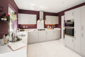 A modern kitchen with light gray cabinets and a purple accent wall features a white countertop with a cookbook and cocktail shaker in the foreground, and stainless steel appliances and a gas range in the background.
