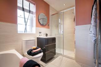 A modern bathroom features a toilet with folded towels in the foreground and a glass-enclosed shower with a vanity and a round mirror on a coral-colored wall in the background.