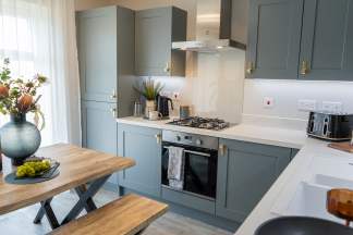A rustic wooden dining table with a vase of flowers sits in the foreground, while a modern grey kitchen with stainless steel appliances and white countertops occupies the background.