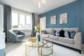 A plush gray sofa with decorative pillows sits in front of a blue accent wall with framed art, while a round armchair and a glass coffee table with a gold accent are in the foreground.