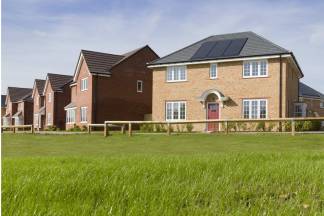 A lush green lawn stretches before a row of modern brick houses, some with solar panels on their roofs, under a clear blue sky.