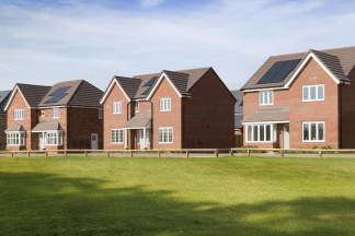 A lush green lawn with a wooden fence in the foreground is in front of a row of modern brick houses with solar panels on their roofs under a clear blue sky.