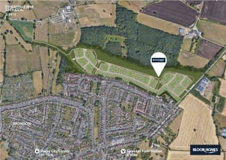A new housing development is being built on a green field adjacent to an existing residential area and a wooded area, with a road and fields visible in the distance.