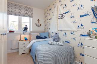 A nautical-themed bedroom features a striped bed with whale pillows in the foreground and a wall covered in sailboat wallpaper in the background.