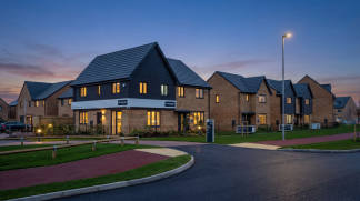 A residential development with modern, brick-built houses featuring slanted roofs and illuminated windows, set against a backdrop of a clear evening sky with a streetlight in the foreground.
