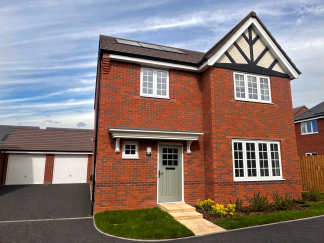 A red brick house with white-framed windows and a light green door stands on a paved driveway in front of a cloudy blue sky, with a detached garage visible to the left.