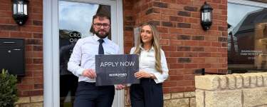 A man and a woman stand in front of a brick building, the man holding a sign that says "APPLY NOW" while the woman stands beside him.