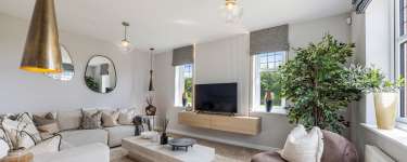 A large sectional sofa and a modern coffee table fill the foreground of a living room, while a television mounted on a wooden cabinet and windows overlooking greenery are in the background.