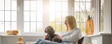 A woman sits with her black poodle in front of a large window, with decorative vases on the windowsill and trees visible outside.