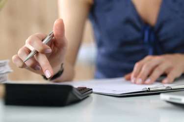 A person's hands are shown in the foreground, one holding a pen over a calculator and the other resting on a clipboard, with a blurred office background.