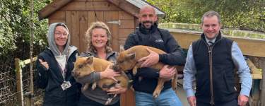The image shows a group of four people, two women and two men, standing in front of a wooden shed and holding two dogs, one of which appears to be a German shepherd or similar breed.