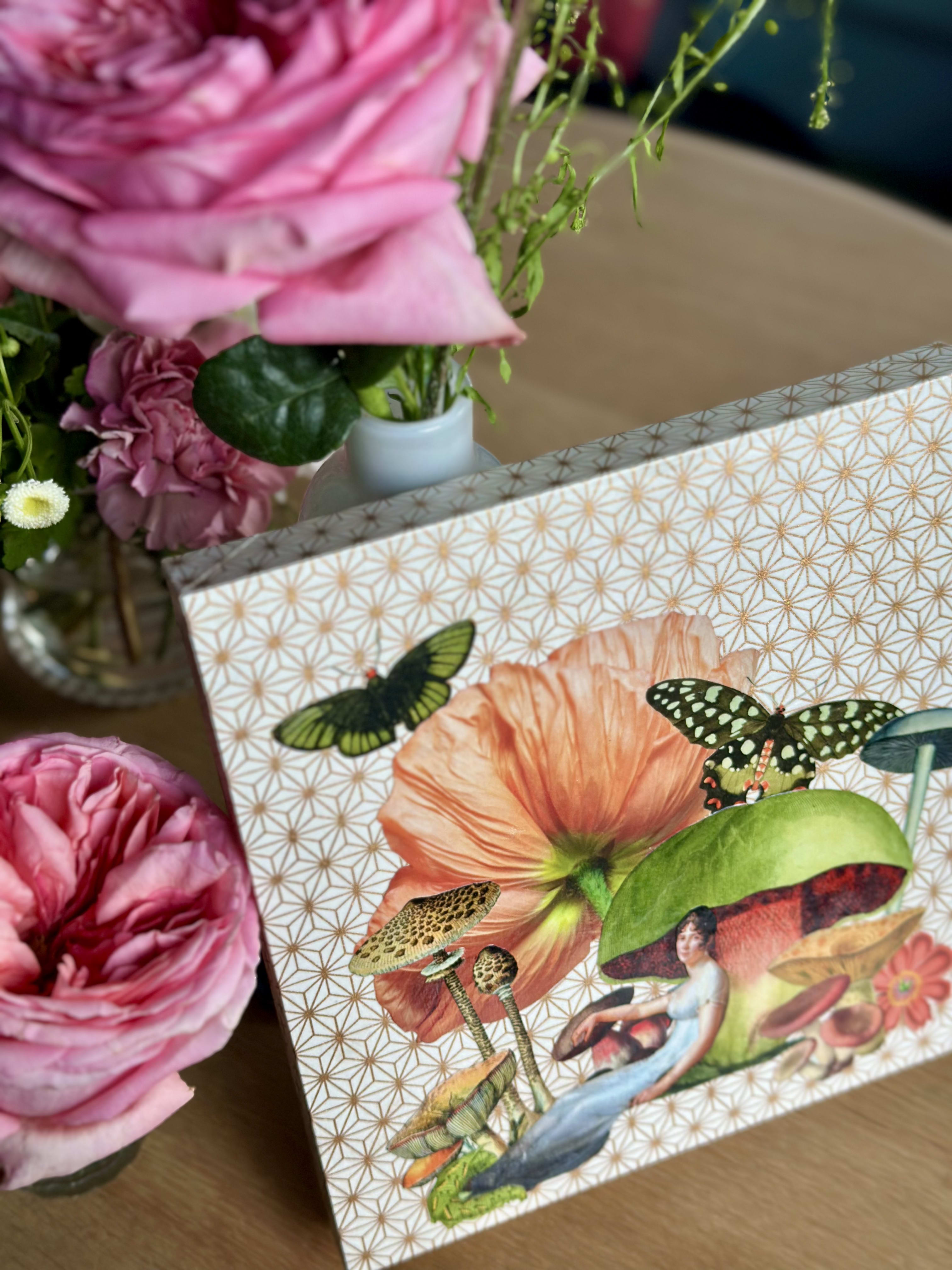 Angled view of the collage resting on a wood table beside pink roses, showing the peach poppy, spotted butterflies, and a woman in a Regency gown reclining beneath a green mushroom cap