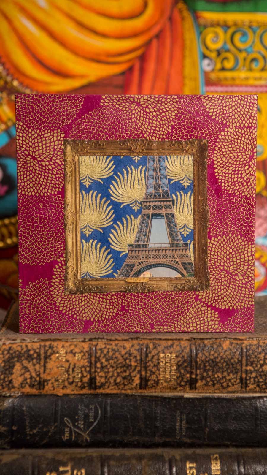 Front view of the collage propped on a stack of antique leather-bound books, its gilt-framed Eiffel Tower centered on magenta paper patterned with gold chrysanthemums