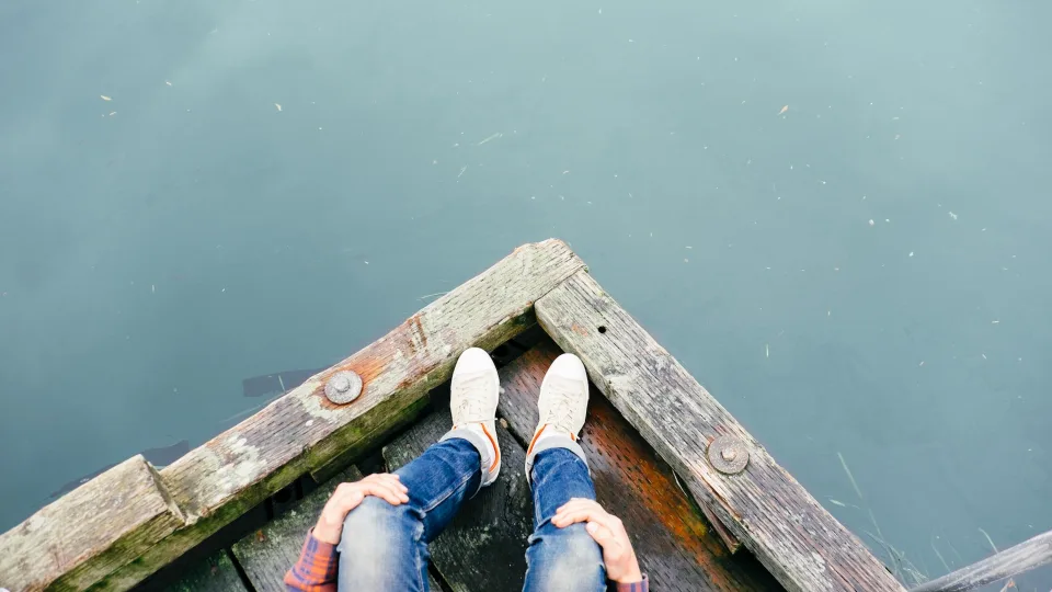 Someone sitting on the dock of a lake wearing jeans and sneakers