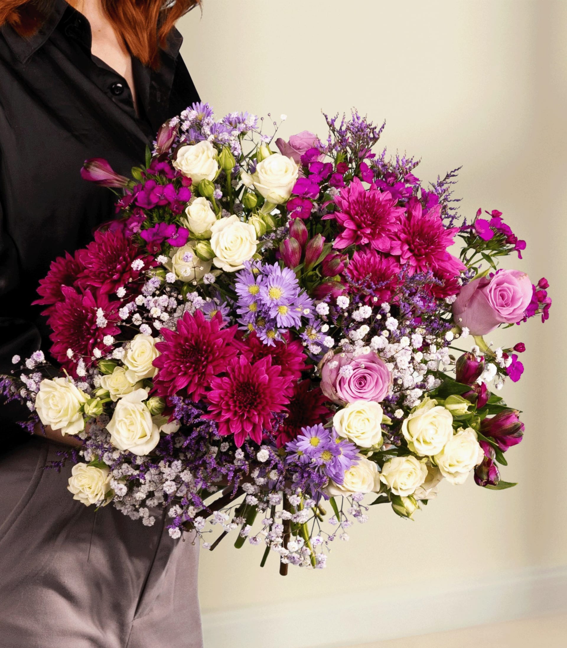 Ein farbenfroher Strauß verschiedener Blumen, darunter rosa Rosen, lila Gänseblümchen und weiße Nelken, gehalten von einer Person in einer schwarzen Jacke vor einem schlichten Hintergrund.