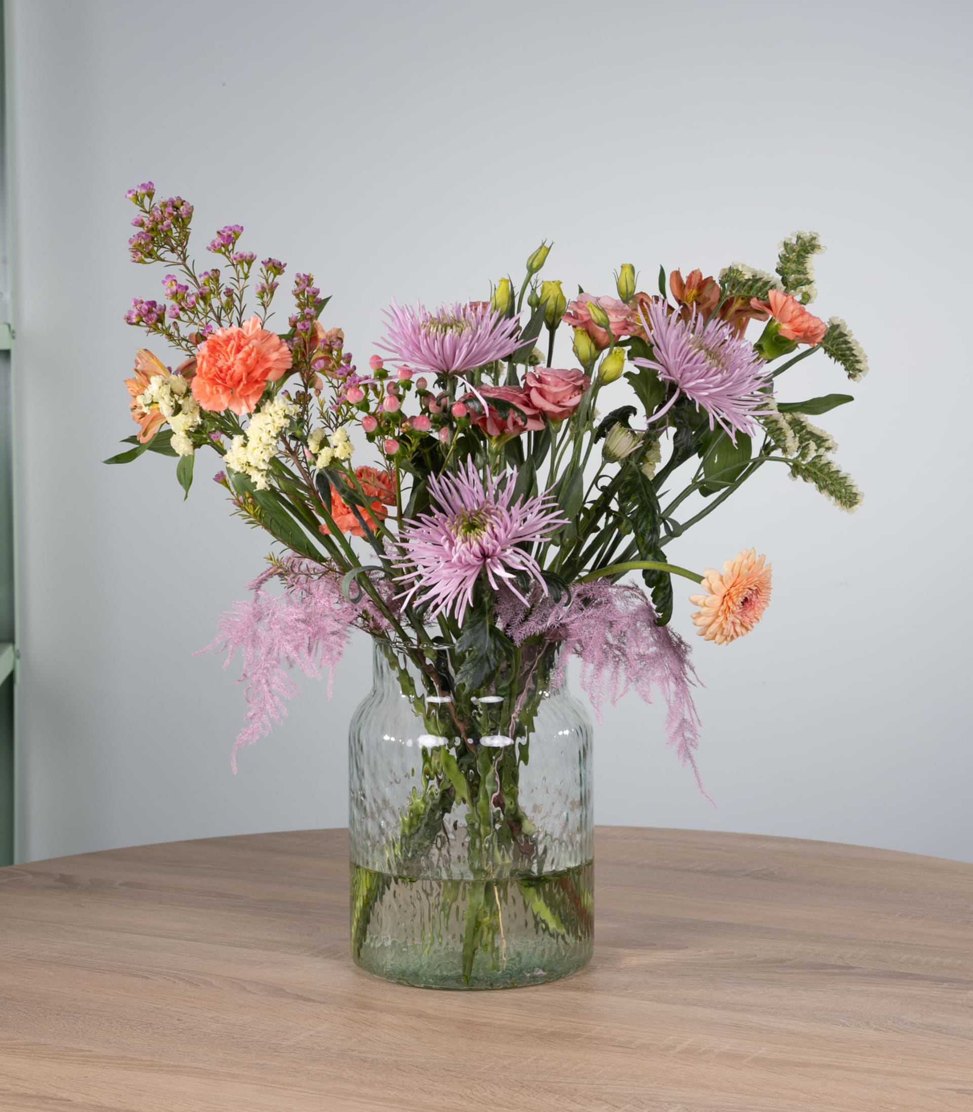 Ein leuchtender Strauß verschiedener bunter Blumen, darunter rosa und orange Dahlien, weiße Gänseblümchen und violette Chrysanthemen, wird in einer Glasvase auf einem Holztisch vor einem schlichten Hintergrund präsentiert.