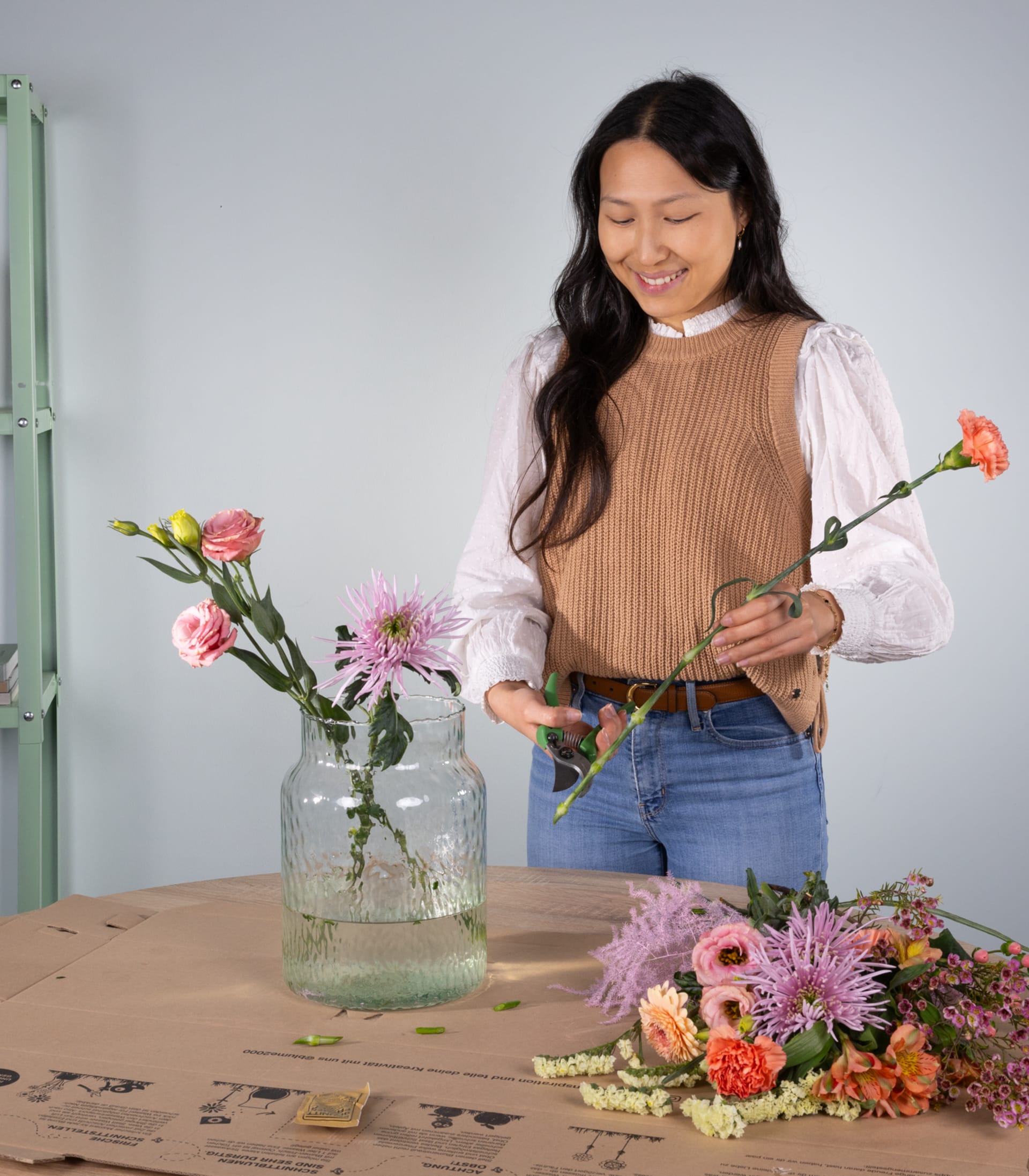 Eine junge Frau mit langem, dunklem Haar arrangiert einen Strauß bunter Blumen auf einem Holztisch. Im Hintergrund stehen ein mit Wasser gefülltes Glas und weitere Blumen.