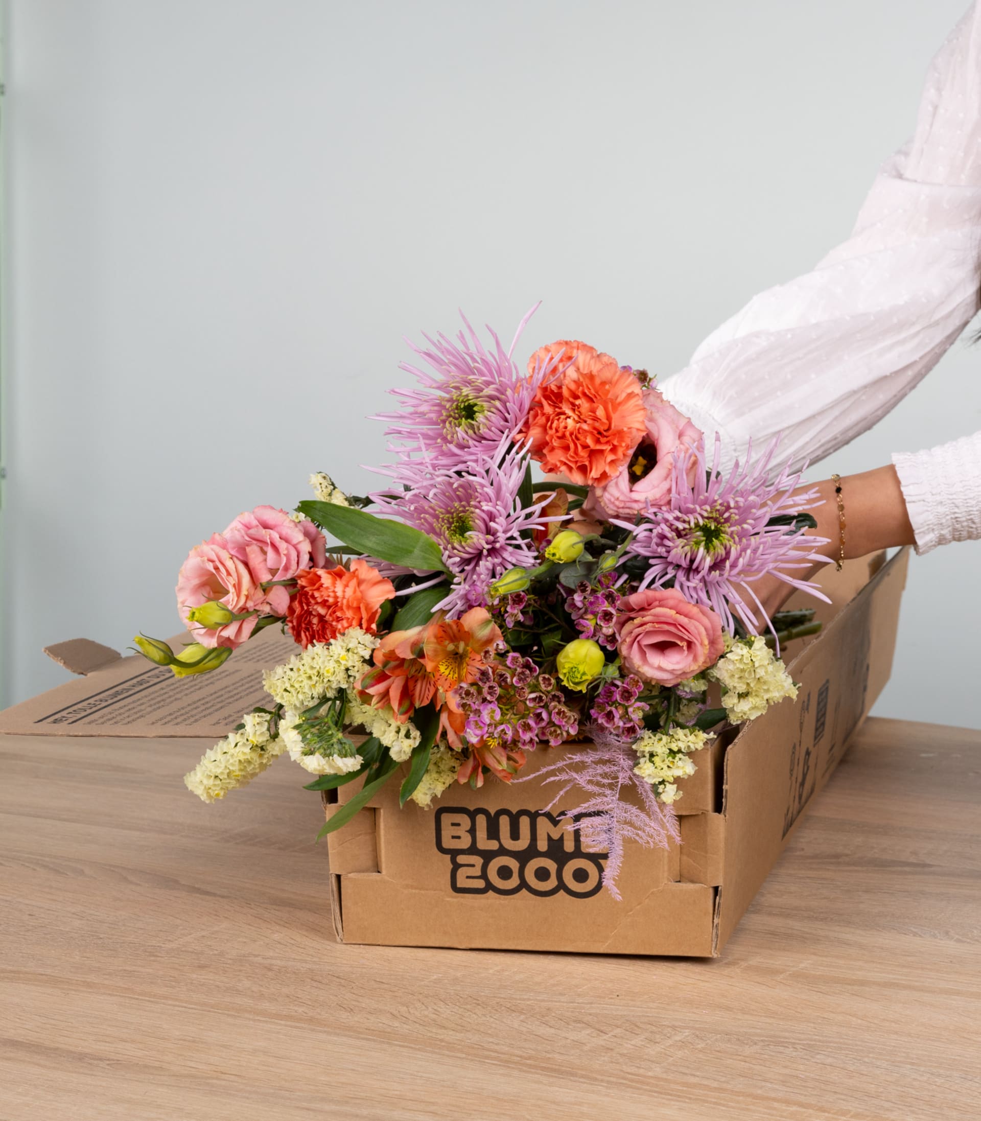 Ein farbenfroher Strauß bunter Blumen, darunter Rosen, Nelken und Chrysanthemen, wird in einem Karton auf einer Holzoberfläche präsentiert.