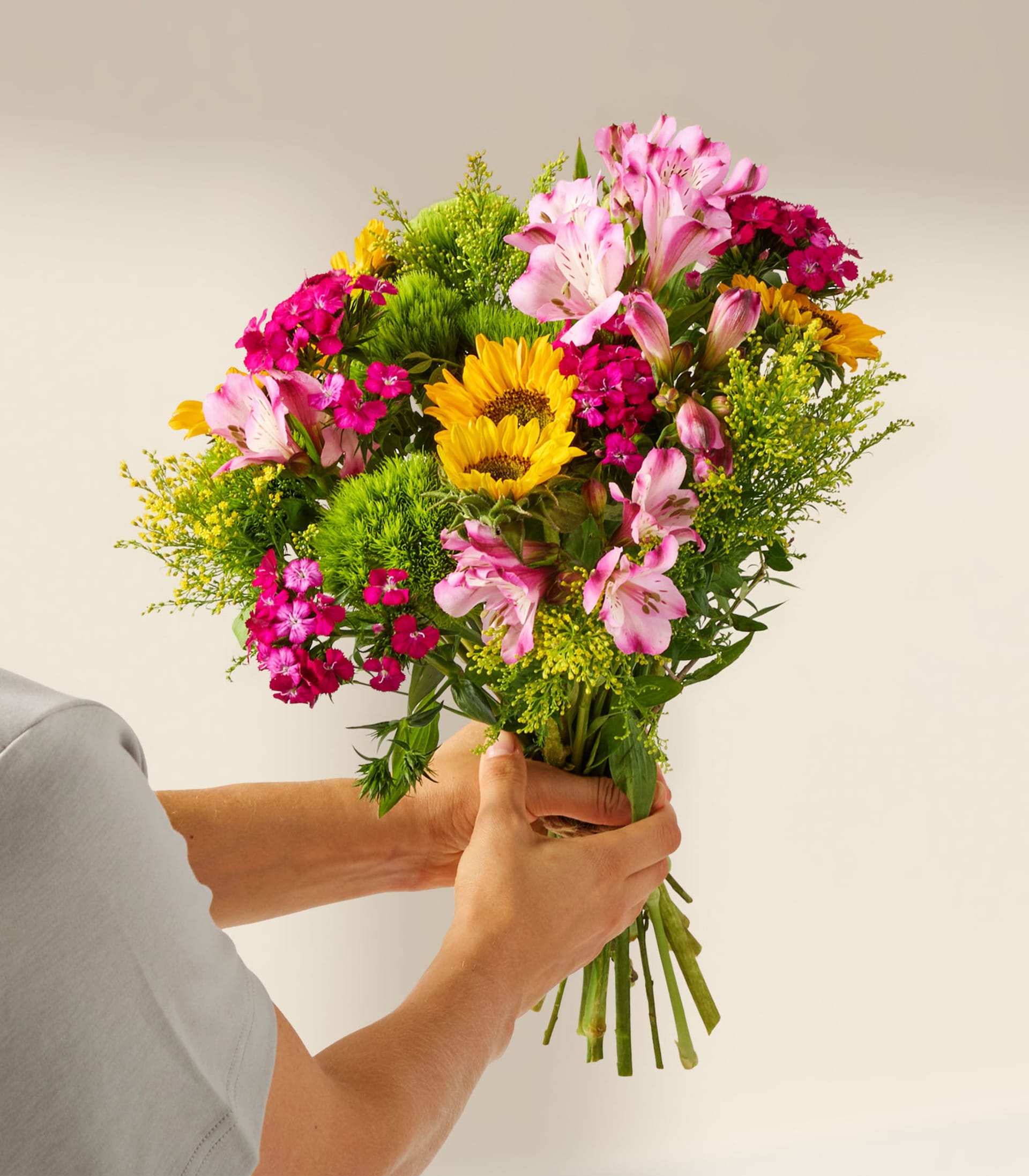 Ein lebendiger Strauß bunter Blumen, darunter Sonnenblumen, rosa Gänseblümchen und grünes Laub, wird in einer Glasvase vor einem schlichten weißen Hintergrund präsentiert.