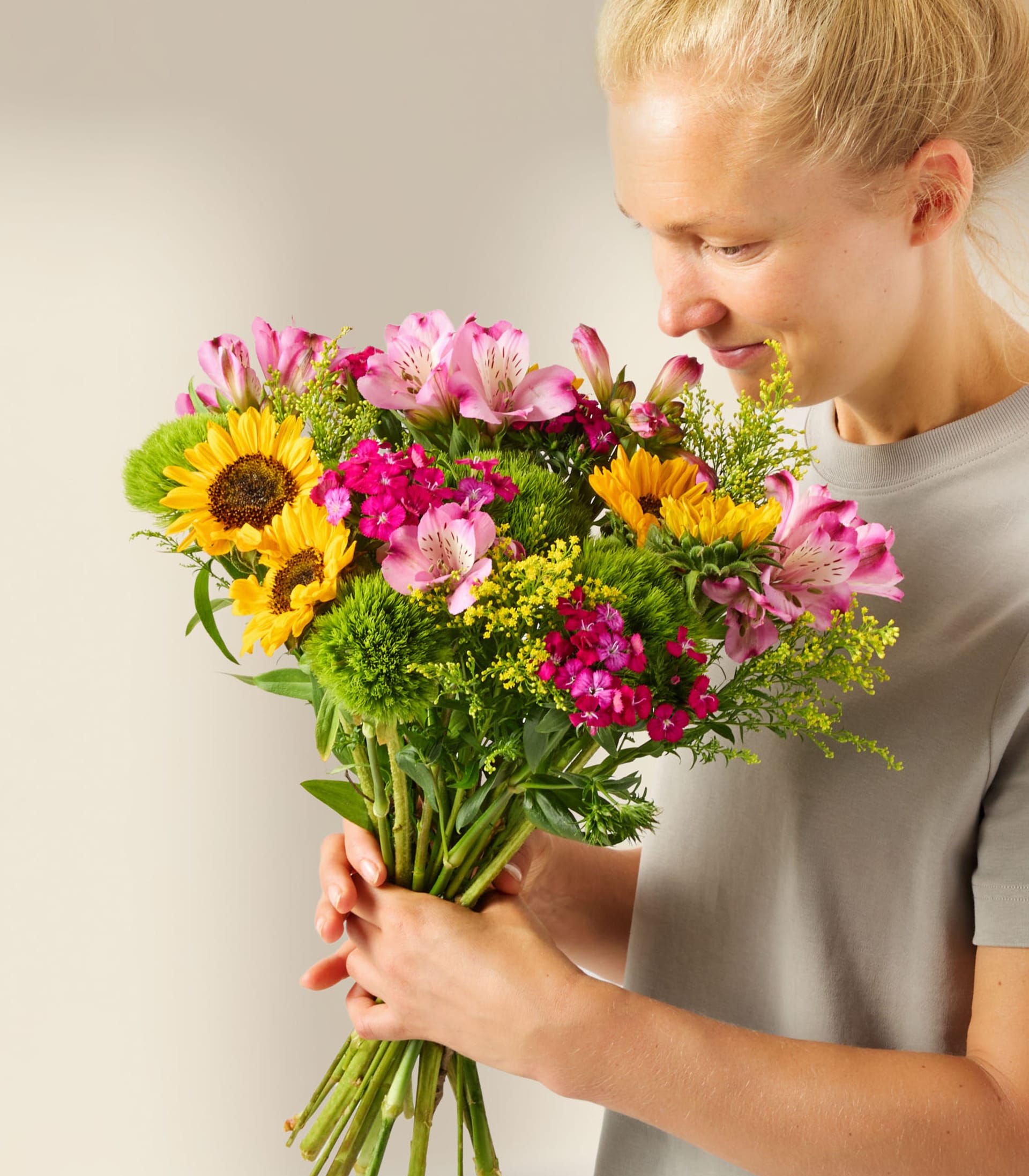 Eine junge Frau hält vor einem schlichten weißen Hintergrund einen leuchtend bunten Blumenstrauß, darunter Sonnenblumen, Nelken und andere Blüten.