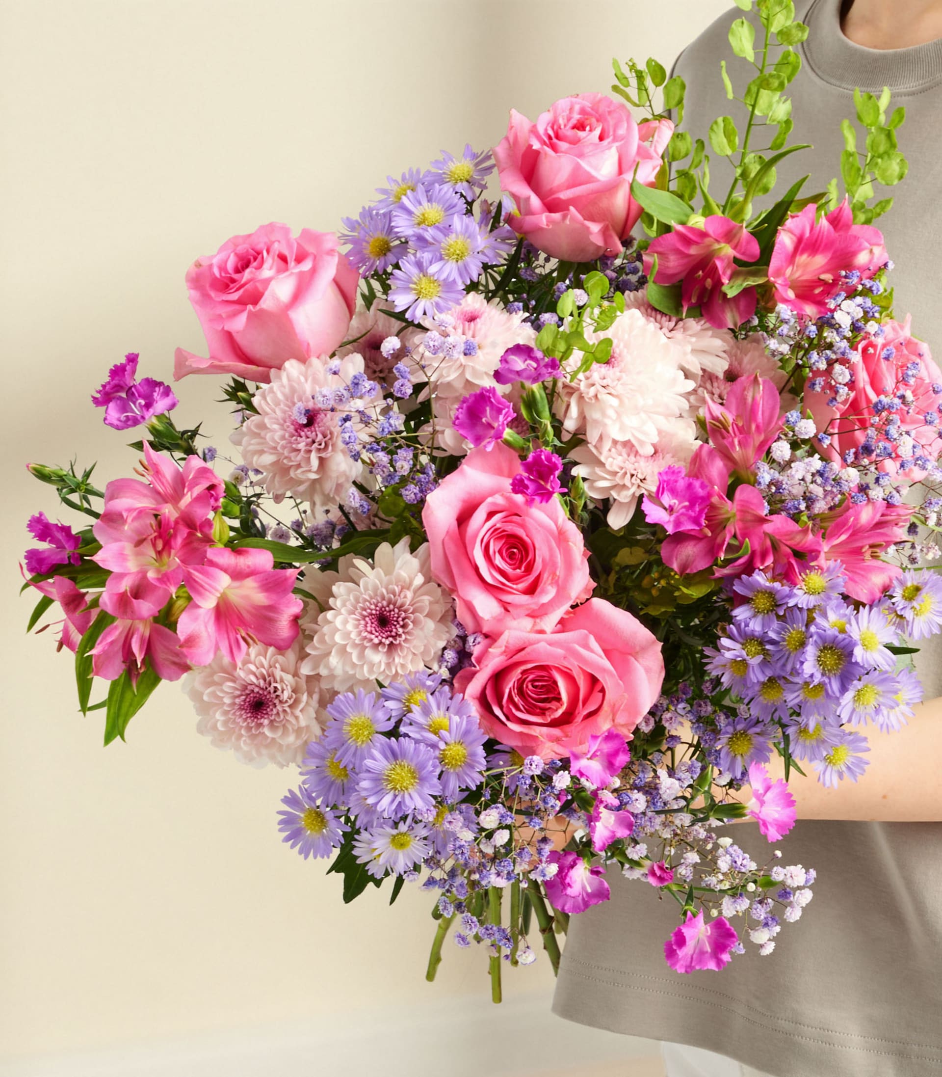 Eine Person hält einen leuchtenden Strauß rosa Rosen, lila Gänseblümchen und andere bunte Blumen vor einem schlichten weißen Hintergrund.