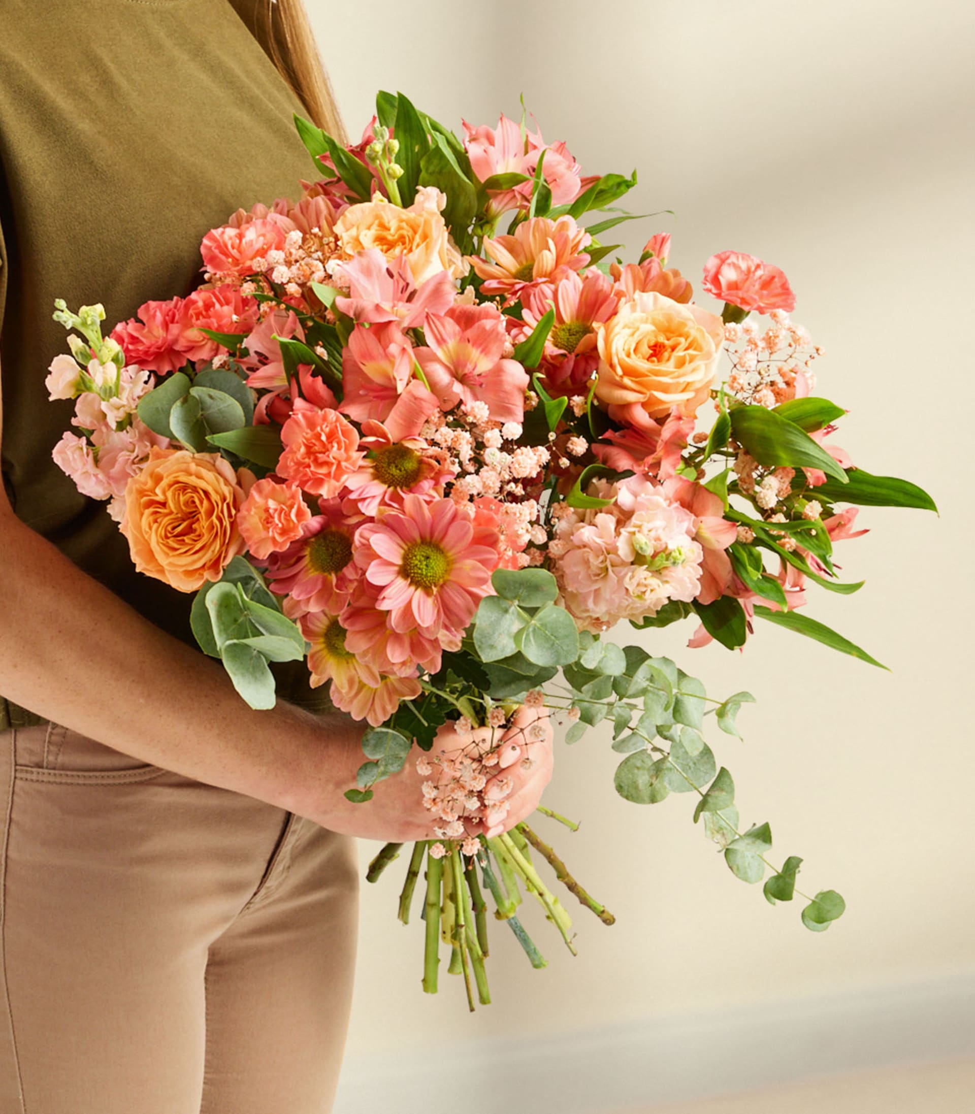 Ein leuchtender Strauß bunter Blumen, darunter Rosen, Nelken und Gänseblümchen, wird vor einem schlichten weißen Hintergrund vor eine Person mit langem, blondem Haar gehalten.