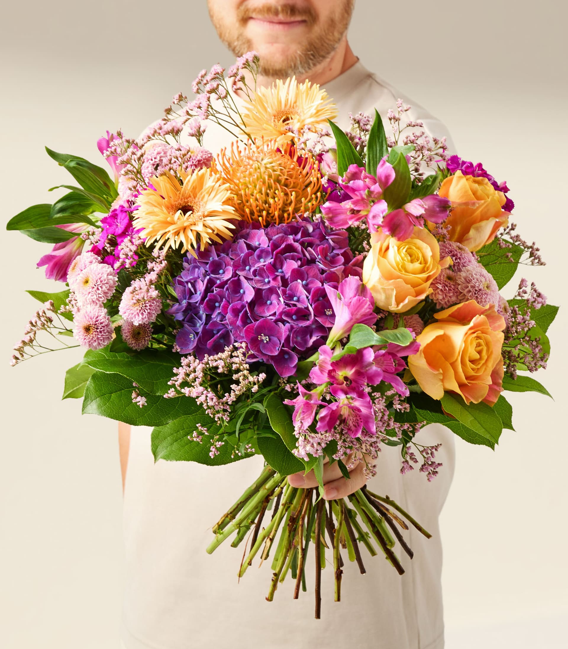 Ein lebendiger Strauß bunter Blumen, darunter Rosen, Gerbera und andere Blüten, wird in einer Glasvase vor einem schlichten weißen Hintergrund präsentiert.