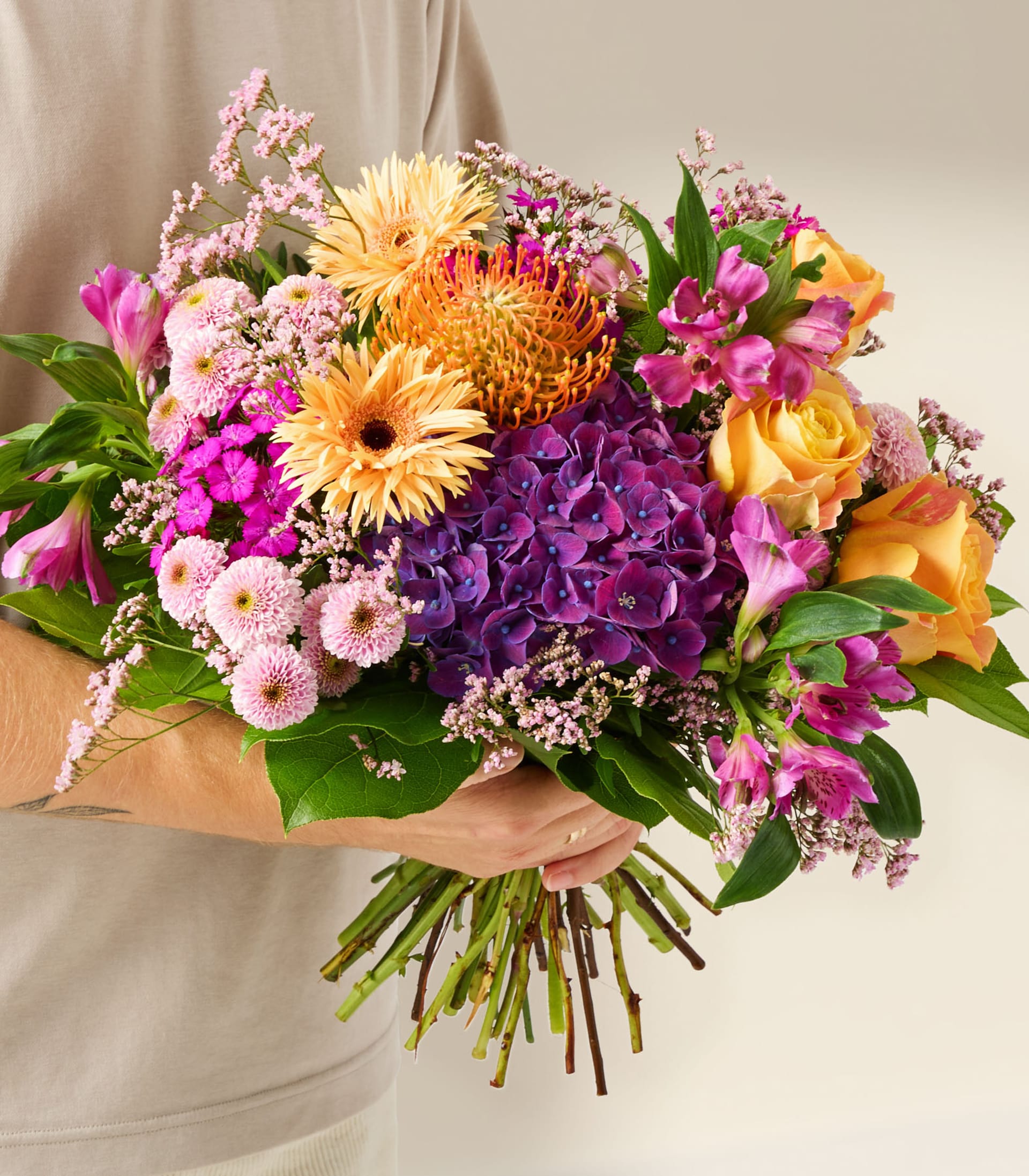 Ein leuchtender Strauß bunter Blumen, darunter gelbe und orangefarbene Gerbera, violette Hortensien sowie rosa und gelbe Rosen, gehalten von einer Person in einem hellen Hemd vor einem schlichten weißen Hintergrund.