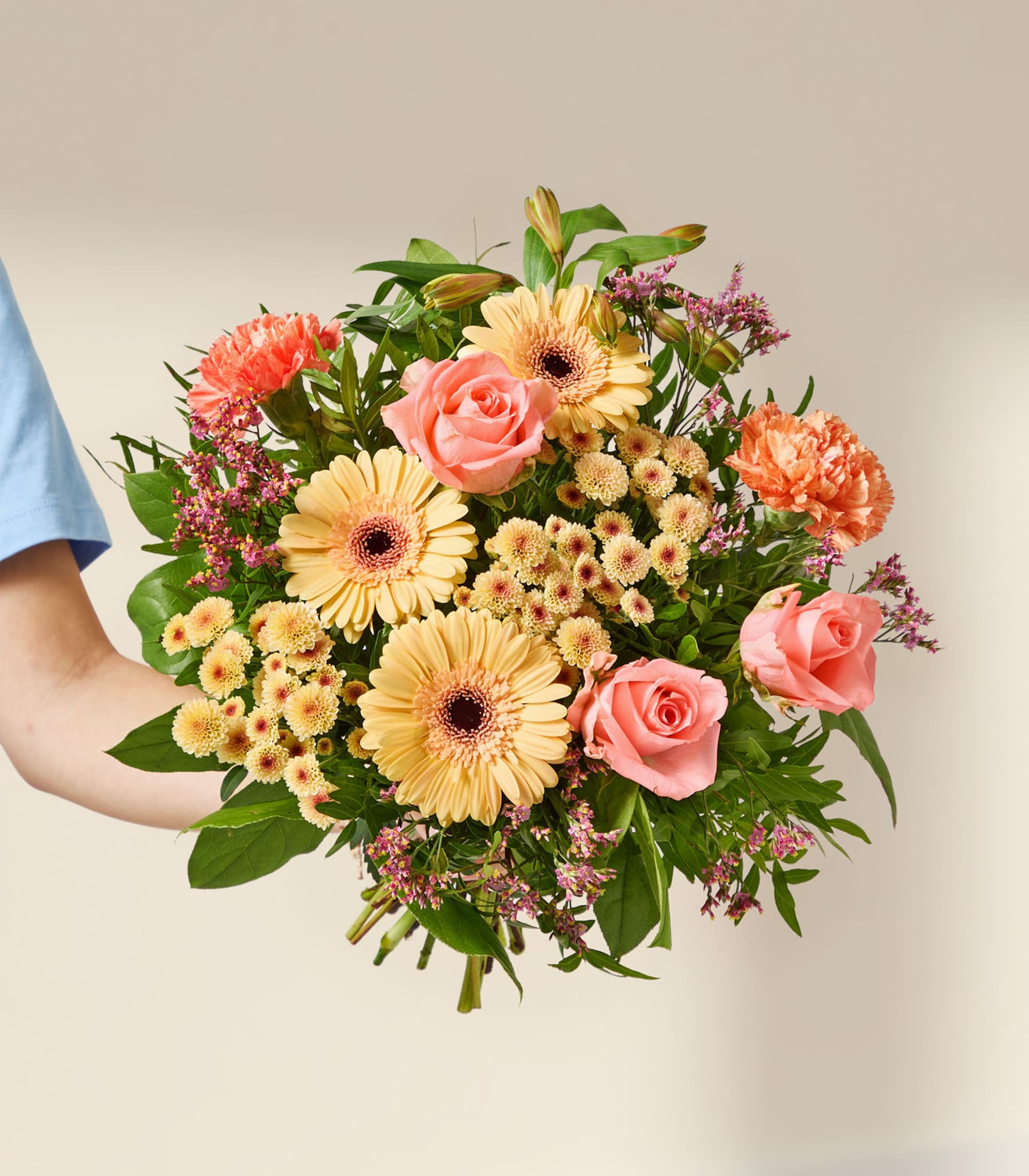 Eine Hand hält einen leuchtenden Strauß aus rosa Rosen, orangefarbenen Nelken und gelben Gerbera vor einem sanften, neutralen Hintergrund.