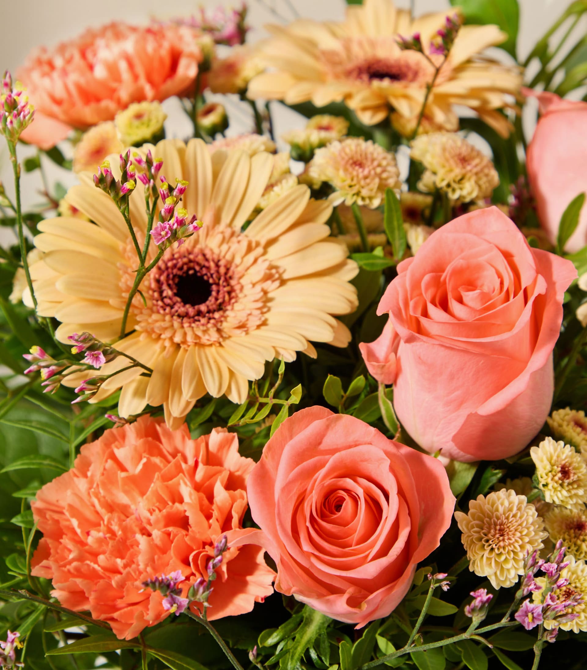 Ein leuchtender Strauß aus pfirsich- und orangefarbenen Blüten, darunter Rosen, Gerbera und Nelken, ist mit üppigem Grün vor einem weichen, verschwommenen Hintergrund arrangiert.