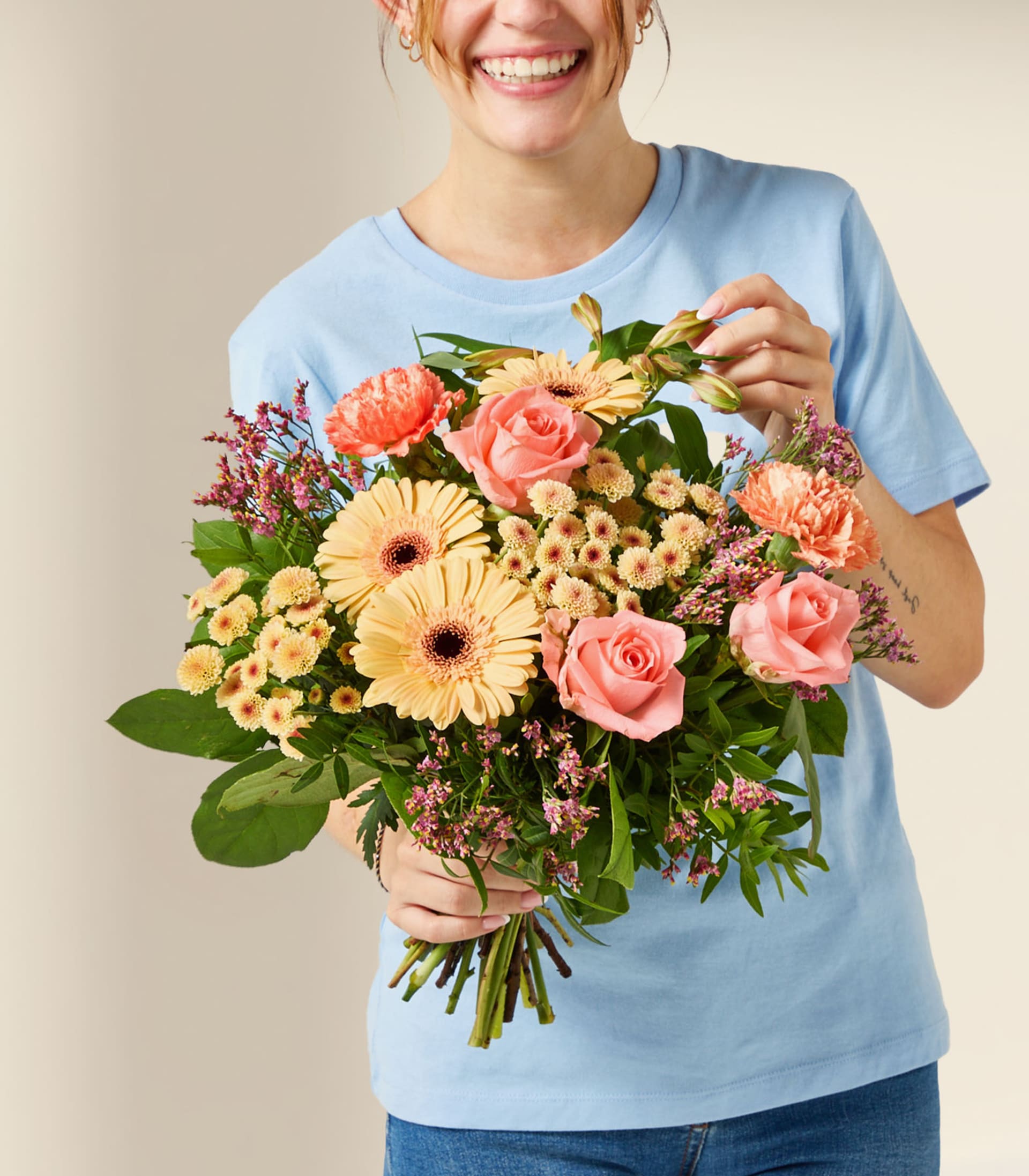 Eine lächelnde Person in einem blauen T-Shirt hält einen farbenfrohen Strauß aus rosa Rosen, pfirsichfarbenen Gerbera und kleinen lila Blüten vor einem schlichten, hellen Hintergrund.