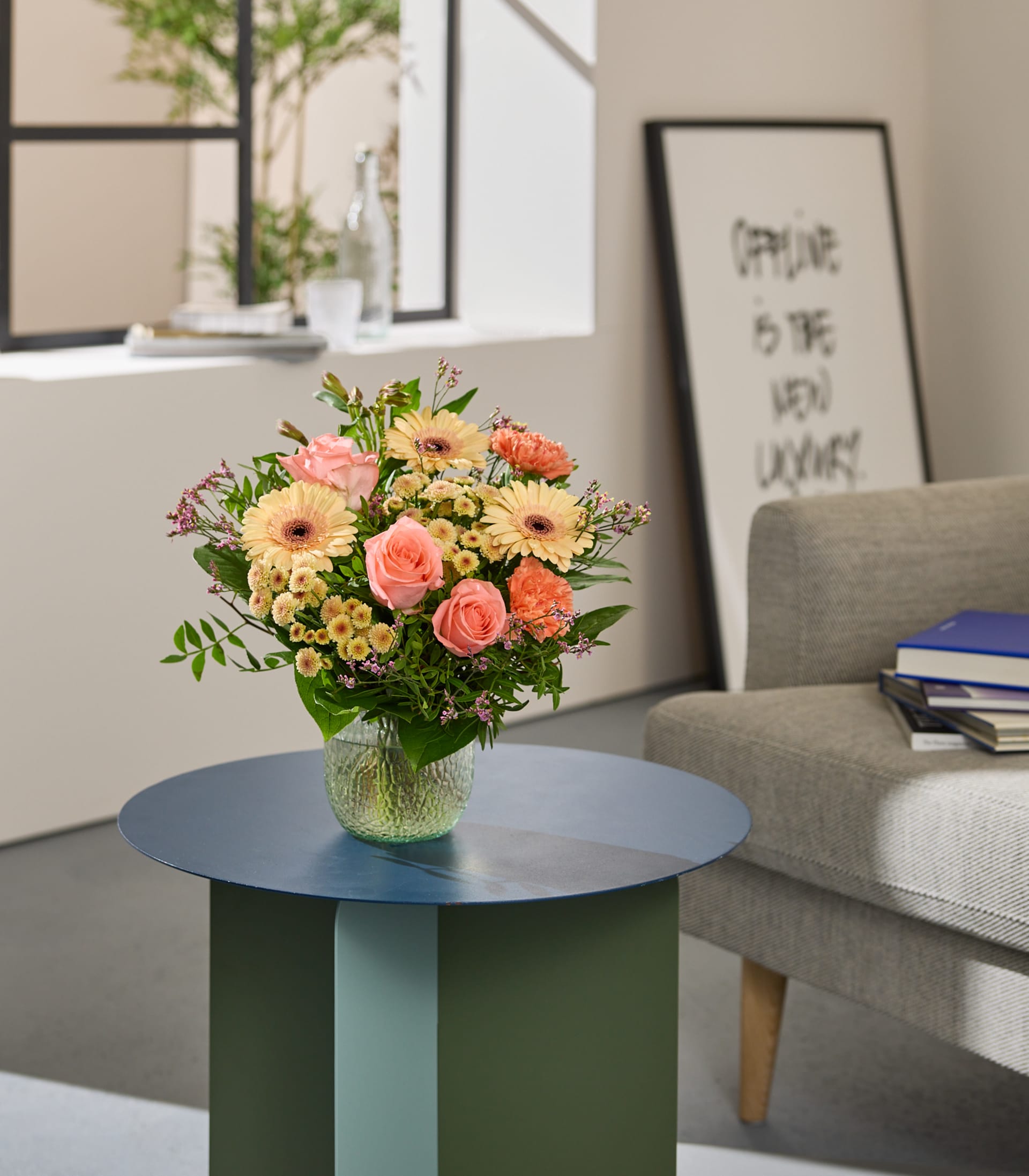 Auf einem runden Tisch in einem gemütlichen Wohnzimmer steht ein farbenfroher Blumenstrauß; im Hintergrund sind ein gerahmtes Kunstwerk und Bücher zu sehen.