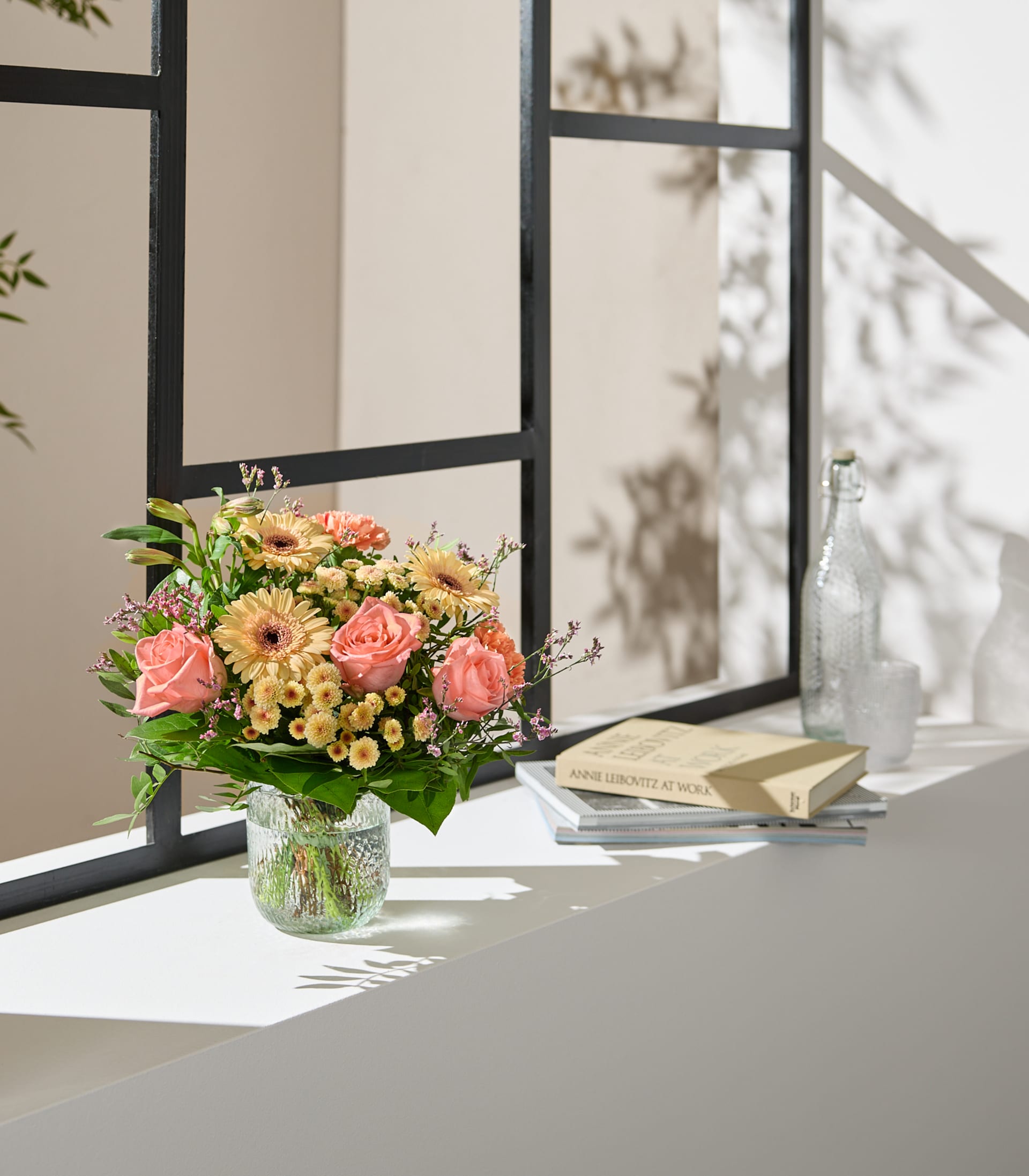 Auf einem Fensterbrett steht eine Vase mit farbenprächtigen Blumen, im Hintergrund ein Buch und andere Gegenstände – eine gemütliche und einladende Szene.