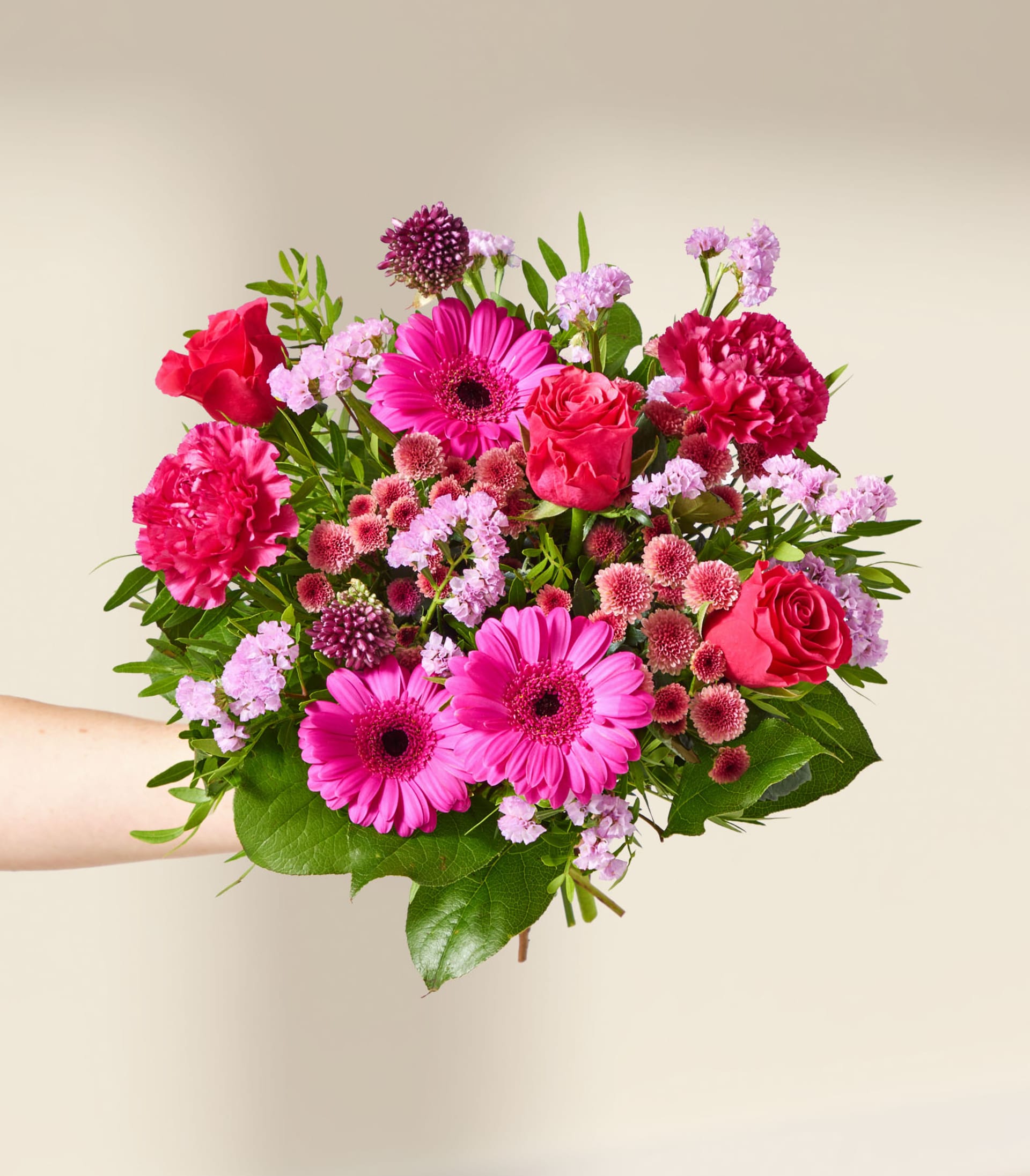 Eine Hand hält einen leuchtenden Strauß rosa und roter Blumen, darunter Gerbera und Rosen, vor einem sanften, neutralen Hintergrund.